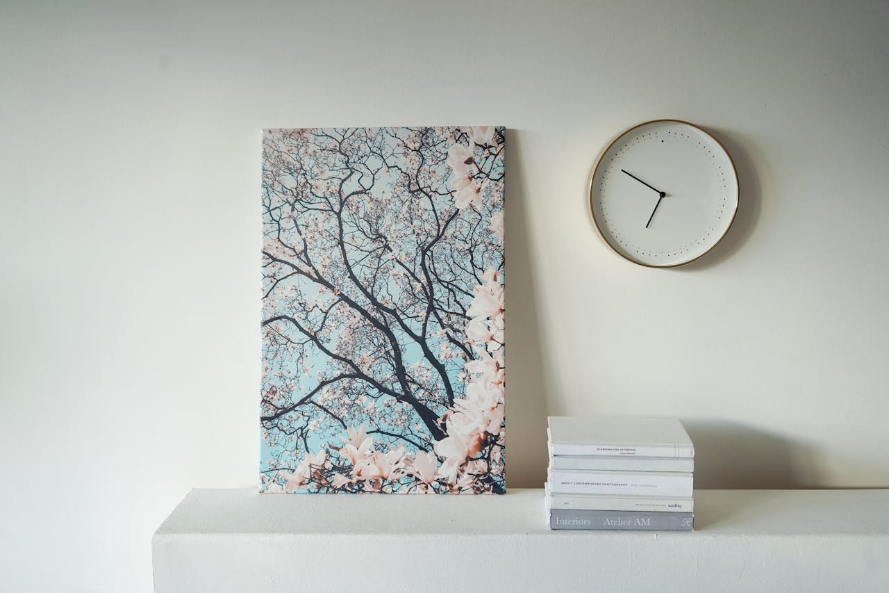 A serene still life featuring a tree painting, clock, and books on a white table.